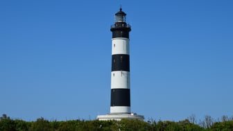 Bodie Island Lighthouse, painted in horizontal black-and-white bands, rises above golden marsh grasses at sunset, with a wooden boardwalk leading toward it across the Outer Banks of North Carolina.